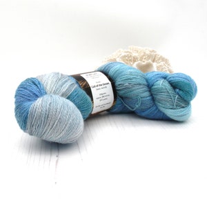 May include: A skein of blue and light blue yarn with a label that reads "Call of the Ocean". The yarn is hand-dyed and appears soft. A white, textured decorative item is in the background.