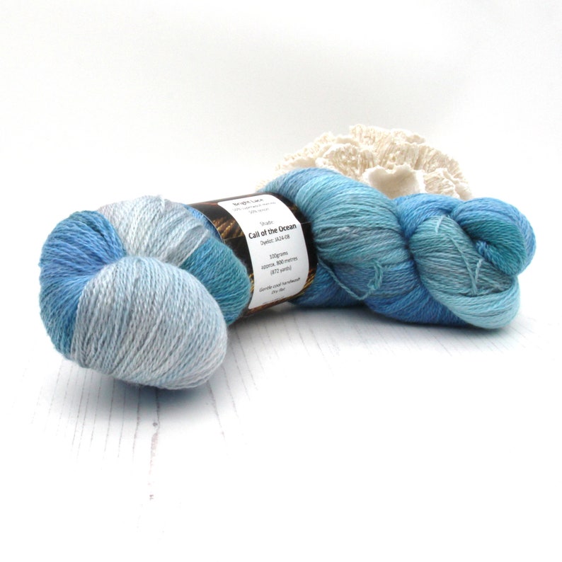 May include: A skein of blue and light blue yarn with a label that reads "Call of the Ocean". The yarn is hand-dyed and appears soft. A white, textured decorative item is in the background.
