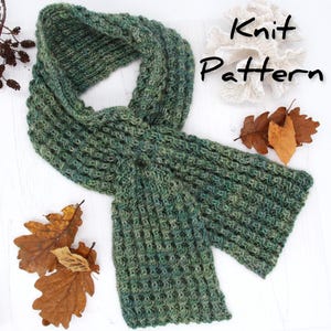 May include: A green and gray knit scarf with a textured pattern. The scarf is displayed on a white surface with dried leaves and the words "Knit Pattern". The scarf is a cozy accessory.