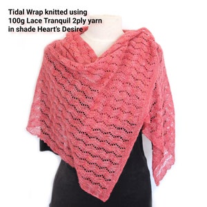 May include: A coral-colored Tidal Wrap shawl, knitted with a lace pattern. The shawl is draped over a mannequin, showcasing its intricate design and texture. The text on the image reads: "Tidal Wrap knitted using 100g Lace Tranquil 2ply yarn in shade Heart's Desire."