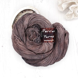 May include: A skein of hand-dyed yarn in shades of brown, purple, and grey. The yarn is wound into a ball and is labeled "Perrans Yarns" with the website address "perranyarns.co.uk".