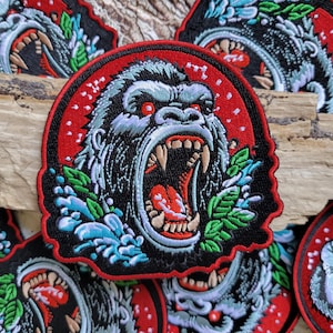 May include: A black and white embroidered patch of a roaring gorilla with red eyes, surrounded by blue and green water and foliage. The patch has a red background with white polka dots.
