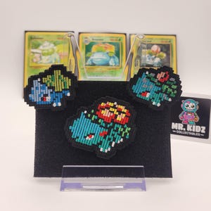 May include: Three embroidered pixel art patches of the Pokemon Bulbasaur. The patches are green, blue, and red with black outlines. The patches are displayed on a black background with a white stand.