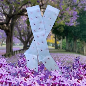 Può includere: Un paio di calze bianche alte fino al ginocchio con un motivo floreale rosa. Le calze sono su un letto di fiori viola.