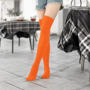 Peut inclure: Chaussettes hautes de couleur orange vif. Les chaussettes sont d'une couleur orange unie et vive et s'étendent des pieds jusqu'au haut des cuisses. Les chaussettes sont portées avec un short en jean. L'arrière-plan est un cadre intérieur flou.