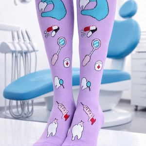 Peut inclure: Paire de chaussettes hautes lavande avec des illustrations sur le thème dentaire. Les chaussettes présentent des images d'une main gantée tenant une dent, des pilules, des miroirs dentaires, une croix rouge, une dent et une seringue. Les chaussettes sont présentées dans un cabinet dentaire.