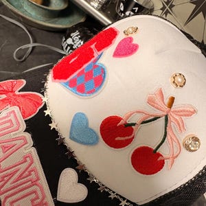 May include: White trucker hat with black mesh backing, adorned with embroidered cherries, hearts, and a bow. Features a chain of stars and decorative embellishments. The hat has the word "DANCE" embroidered on the side.