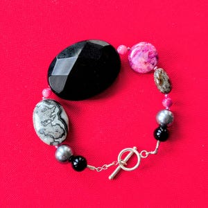 Peut inclure: Un bracelet de perles avec une grosse pierre noire facettée, une pierre marbrée grise et blanche, et diverses autres perles colorées. Le bracelet a un fermoir à bascule argenté sur fond rouge.