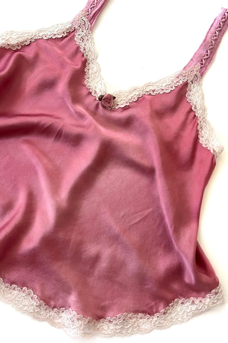 May include: A pink satin camisole with white lace trim and a rose-shaped button at the neckline.