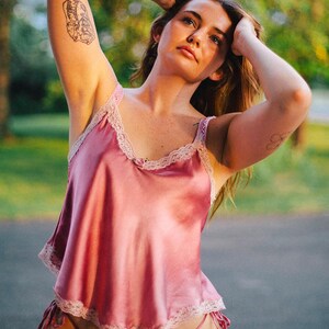 May include: A woman wearing a pink satin and lace lingerie set. The top is a sleeveless cami with a lace trim and the bottom is a high-cut panty with a lace trim and ties at the sides.