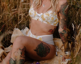 May include: A woman in a yellow floral lingerie set and a straw hat, seated in long grass. The lingerie has a lace-up front. Tattoos decorate the arms and legs. A bouquet of purple and blue flowers is placed nearby.