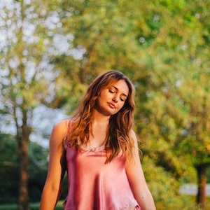 May include: A woman wearing a pink satin top with lace trim and matching pink lace underwear. She is standing in front of a blurred background of green trees.