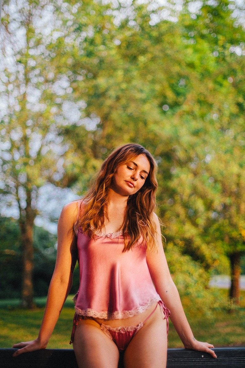 May include: A woman wearing a pink satin top with lace trim and matching pink lace underwear. She is standing in front of a blurred background of green trees.
