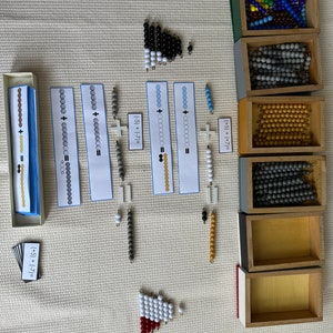 Può includere: Un set di materiali matematici Montessori per imparare l'addizione e la sottrazione. I materiali includono perline di legno di vari colori, carte con equazioni e una cornice di legno a forma di triangolo.