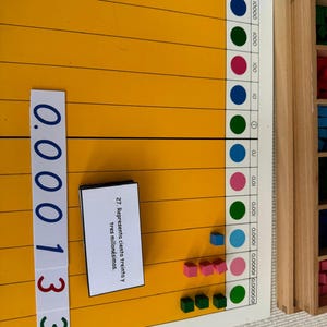 Pode incluir: Um quadro amarelo com um gráfico de números e um cartão que diz "Representa cento e trinta e três milionésimos". Há blocos coloridos no quadro que representam o número 0,000133.