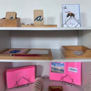 Puede incluir: Una estantería blanca con tres estantes. El estante superior tiene una caja de madera con la letra "a" en ella, una letra "a" de madera en un soporte y una tarjeta con una abeja y la palabra "abeja" en ella. El estante del medio tiene una bandeja de madera con un círculo azul y un lápiz rojo. El estante inferior tiene una pila de papel rosa, una estera de tela enrollada, una bandeja de madera con letras rojas y azules y una tarjeta rosa con la palabra "mar" en ella.