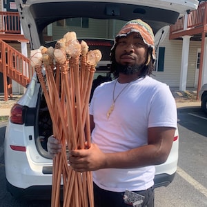 May include: A bundle of copper rods topped with clear quartz crystals, held by a person. The crystals are wrapped with copper wire. The rods are a reddish-brown color. The person is wearing a white t-shirt and a colorful hat.