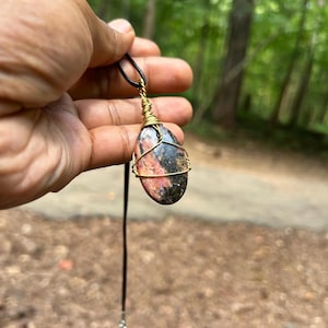 May include: A handcrafted pendant necklace featuring a polished, oval-shaped stone with pink, black, and gold tones. The stone is wrapped in gold-colored wire and suspended from a black cord necklace with a silver-toned chain.