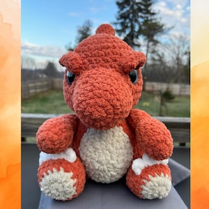 Puede incluir: Un dinosaurio de peluche de crochet de color naranja con detalles blancos. El dinosaurio está sentado con las piernas cruzadas y tiene una expresión de enfado.