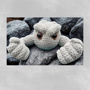 May include: A crocheted gray creature with large brown eyes and a stern expression. The handmade plush toy has a textured, rock-like appearance, with arms and a head. It is posed on a bed of gray rocks, creating a natural setting.