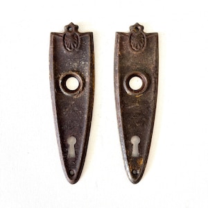 May include: Two matching dark brown, weathered metal door plates with a tapered, elongated shape. Each plate features a circular knob hole at the top, a keyhole opening at the bottom, and a small decorative emblem at the very top edge.