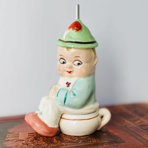 Könnte beinhalten: Vintage Keramikfigur eines sitzenden Kindes mit grünem Hut mit rotem Akzent und hellblauer Jacke. Die Figur sitzt auf einer weißen Teetasse mit Henkel und hat einen silbernen Metallausguss.