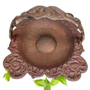 May include: A detailed overhead shot of a decorative, antique-style, cast iron bowl. The bowl is a rusty brown color with ornate floral and scrollwork around the rim and base. The bowl has a central, raised circular design. Green leaves are at the base.