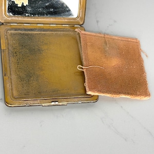 May include: Open antique compact with a mirror and a powder compartment. The compact is gold-toned with a rectangular shape. A peach-colored powder puff rests beside the open compact. The compact is on a white surface.