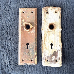 May include: Two rectangular metal door plates with a central circular knob opening and a lower keyhole. The left plate features a weathered, rusted brown finish, while the right plate displays a heavily chipped, distressed white painted surface.