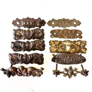 May include: A collection of ten assorted metal drawer pulls and escutcheons in various shapes and finishes. The hardware features ornate scrollwork, floral motifs, and weathered brown or tarnished brass tones, with some pieces showing signs of wear.