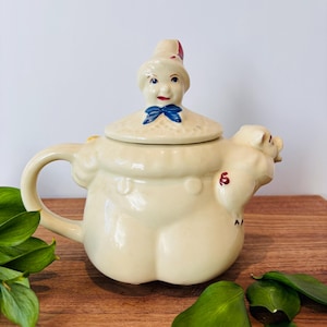 May include: Cream-colored ceramic teapot with a whimsical design. The lid is shaped like a woman's head with a blue bow, and the body of the teapot is shaped like a pig. The number "6" is painted on the side.