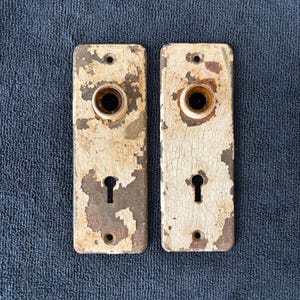 May include: Two rectangular metal door plates with a distressed, cracked white paint finish over a dark, rusted base. Each plate features a central circular opening for a knob, a lower keyhole, and two small screw holes at the top and bottom.