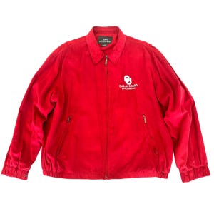 May include: A bright red zip-up jacket with a collar and two front pockets. The jacket features the text "OKLAHOMA. BOOMER" and a logo on the front. The jacket is made of a soft material.