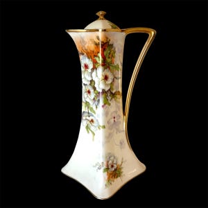 Może przedstawiać: Wysoki dzbanek z porcelany w kolorze kremowym z ręcznie malowanym motywem kwiatowym w kolorze białym i fioletowym, zielonymi liśćmi oraz pomarańczowymi akcentami. Naczynie posiada złocony uchwyt, pasującą pokrywkę ze złotym zwieńczeniem i rozszerzaną podstawę.
