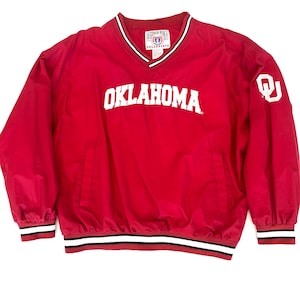 May include: A red Oklahoma Sooners windbreaker jacket with a V-neck collar. The word "OKLAHOMA" is printed in white across the chest. The cuffs and hem have black and white stripes. The Oklahoma "OU" logo is on the right sleeve.