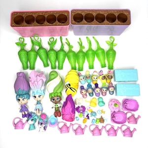 Puede incluir: Una colección de juguetes coloridos, que incluye muñecas con pelo en forma de flor, figuras verdes con forma de planta, regaderas rosas y pequeñas figuras. El conjunto también incluye dos contenedores rectangulares con interiores marrones y varios accesorios pequeños.
