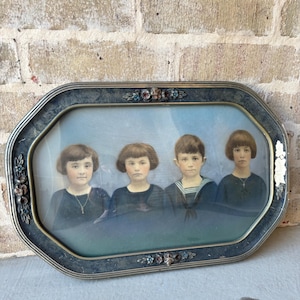 May include: Antique framed portrait featuring four children with short haircuts, dressed in dark clothing. The frame is an octagonal shape with floral accents and a silver finish. The background is a light blue.