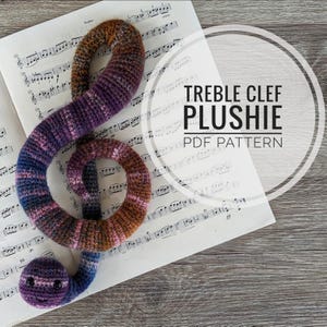 Puede incluir: Un juguete de peluche de clave de sol tejido a crochet en tonos de morado, marrón y rosa, sobre una hoja de música. El texto "TREBLE CLEF PLUSHIE PDF PATTERN" está en un círculo blanco en la hoja de música.