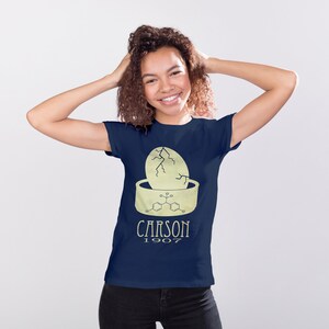 May include: A navy blue t-shirt with a yellow graphic of a cracked egg with a chemical formula and the text "CARSON 1907".
