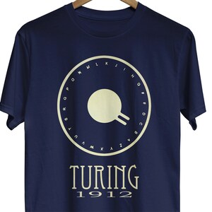 May include: A navy blue t-shirt with a white circle design featuring a stylized symbol and the letters of the alphabet around the perimeter. The text "TURING 1912" is printed below the circle.