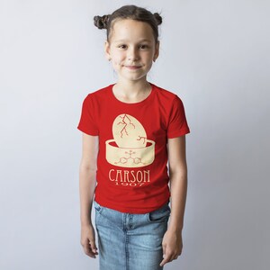 May include: A red t-shirt with a yellow graphic of a cracked egg and a chemical formula. The text "CARSON 1907" is printed below the graphic.