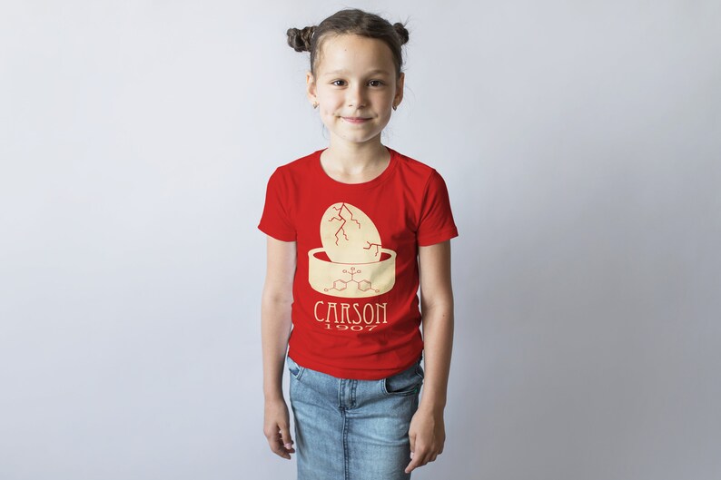May include: A red t-shirt with a yellow graphic of a cracked egg and a chemical formula. The text "CARSON 1907" is printed below the graphic.