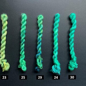 May include: Five skeins of green embroidery floss, each with a different shade of green. The skeins are numbered 23, 25, 29, 24, and 30. The floss is twisted and has a slight sheen, with variations in color from light to dark.