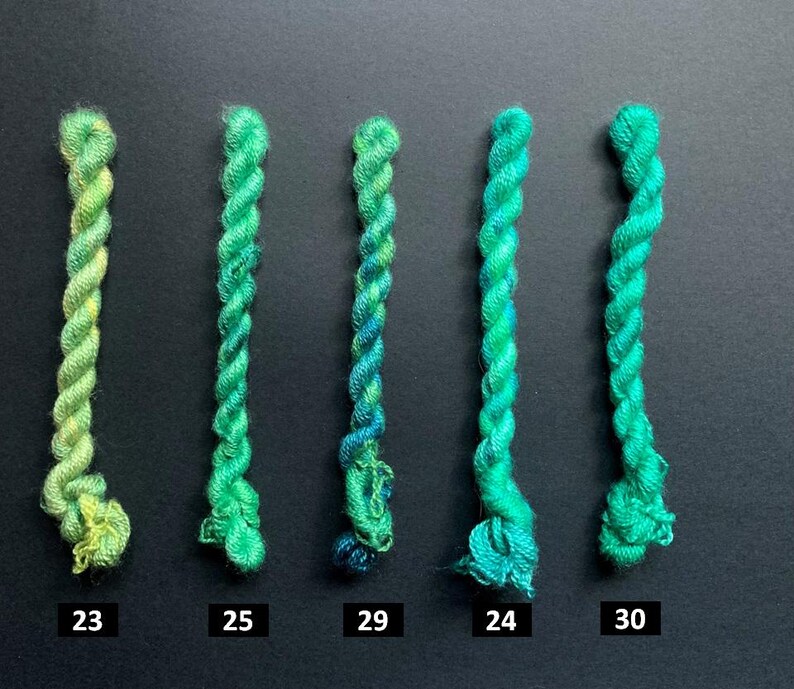 May include: Five skeins of green embroidery floss, each with a different shade of green. The skeins are numbered 23, 25, 29, 24, and 30. The floss is twisted and has a slight sheen, with variations in color from light to dark.