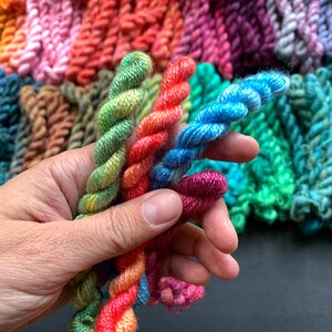 May include: Close-up of colorful yarn skeins in various shades of green, orange, blue, and purple. The yarn is tightly twisted, with a textured appearance. Other skeins of yarn are visible in the background, creating a vibrant display of colors.