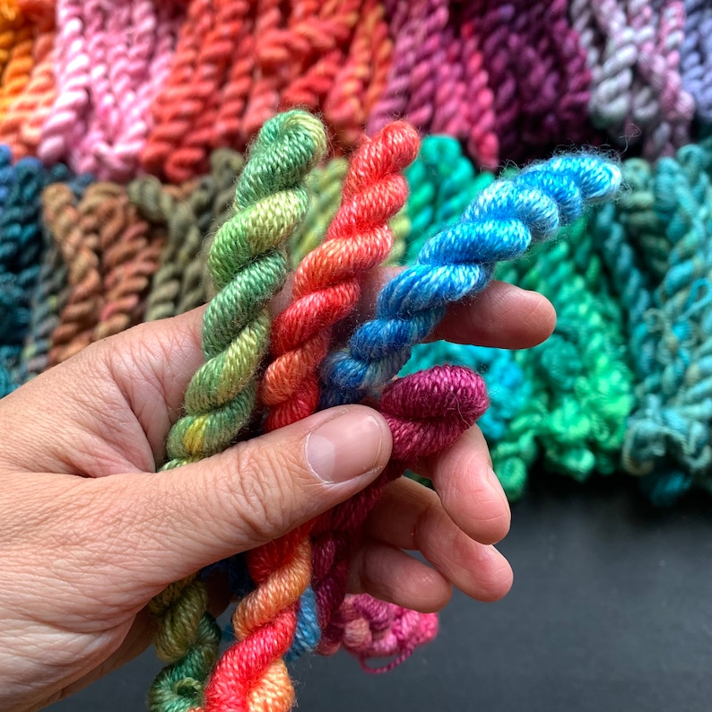 May include: Close-up of colorful yarn skeins in various shades of green, orange, blue, and purple. The yarn is tightly twisted, with a textured appearance. Other skeins of yarn are visible in the background, creating a vibrant display of colors.
