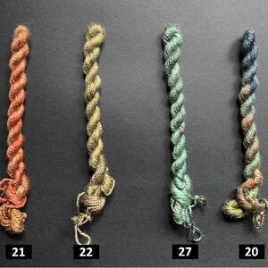 May include: Four twisted embroidery floss skeins in shades of orange, gold, green, and blue. Each skein is labeled with a number: 21, 22, 27, and 20. The floss is tightly wound, with a slightly frayed end on each.