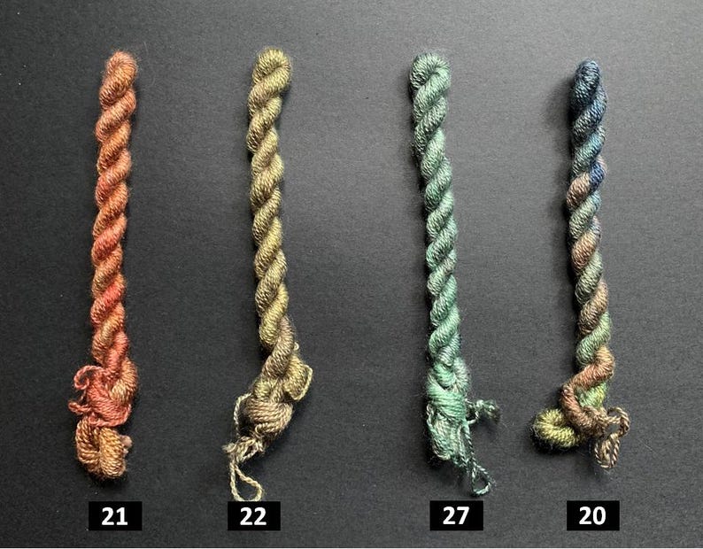 May include: Four twisted embroidery floss skeins in shades of orange, gold, green, and blue. Each skein is labeled with a number: 21, 22, 27, and 20. The floss is tightly wound, with a slightly frayed end on each.