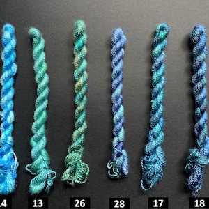 May include: Six skeins of colorful embroidery floss in shades of blue and green are arranged vertically against a dark background. Each skein is labeled with a number. The floss is tightly twisted, showcasing the vibrant color variations.