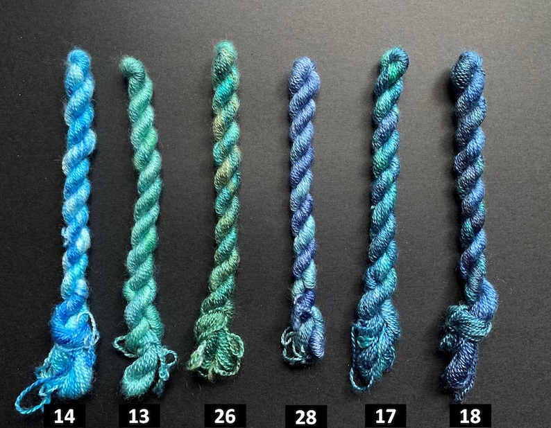 May include: Six skeins of colorful embroidery floss in shades of blue and green are arranged vertically against a dark background. Each skein is labeled with a number. The floss is tightly twisted, showcasing the vibrant color variations.